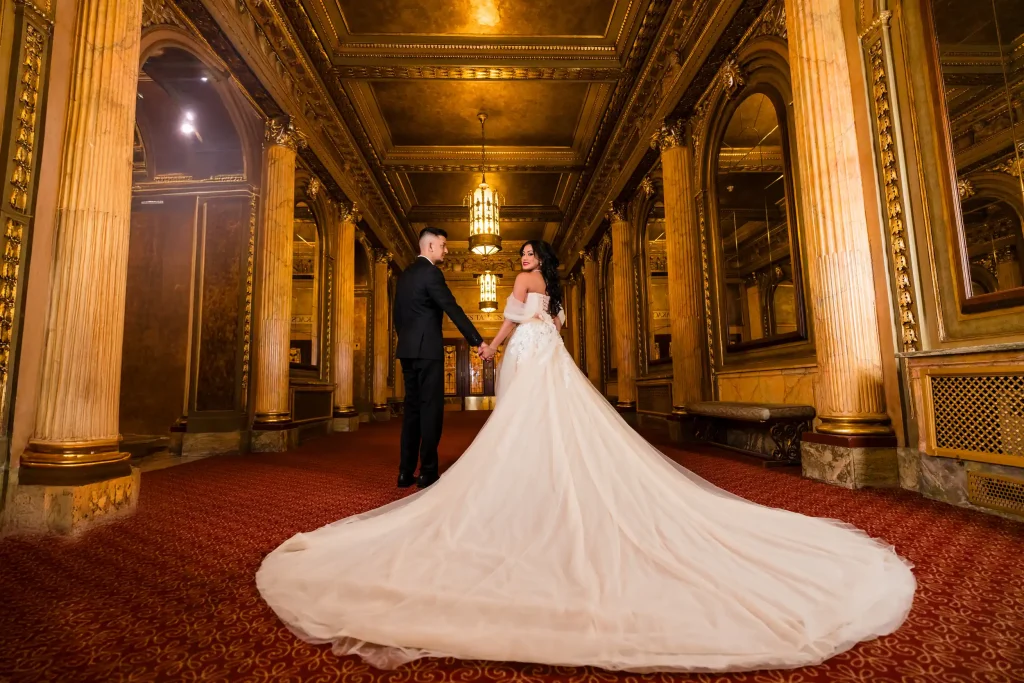 A couple standing in a grand gilded hall, the woman wearing a breathtaking white long-train gown — its train stretching the full length of the corridor behind her. Engagement shoot dress by Athena Dress Rental.