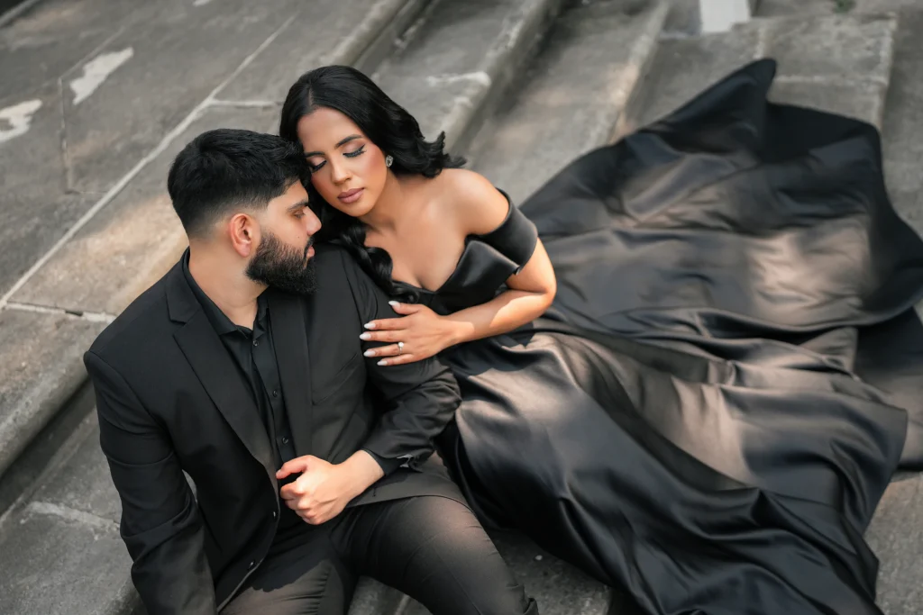 A couple seated on stone steps for an engagement photoshoot, the woman dressed in a sweeping black long-train gown — fabric pooling dramatically around them. Engagement photo outfits from Athena Dress Rental.
