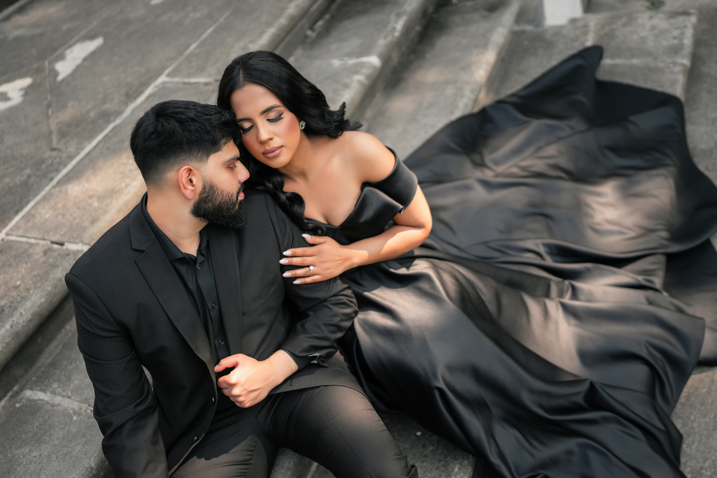 A couple seated on stone steps for an engagement photoshoot, the woman dressed in a sweeping black long-train gown — fabric pooling dramatically around them. Engagement photo outfits from Athena Dress Rental.