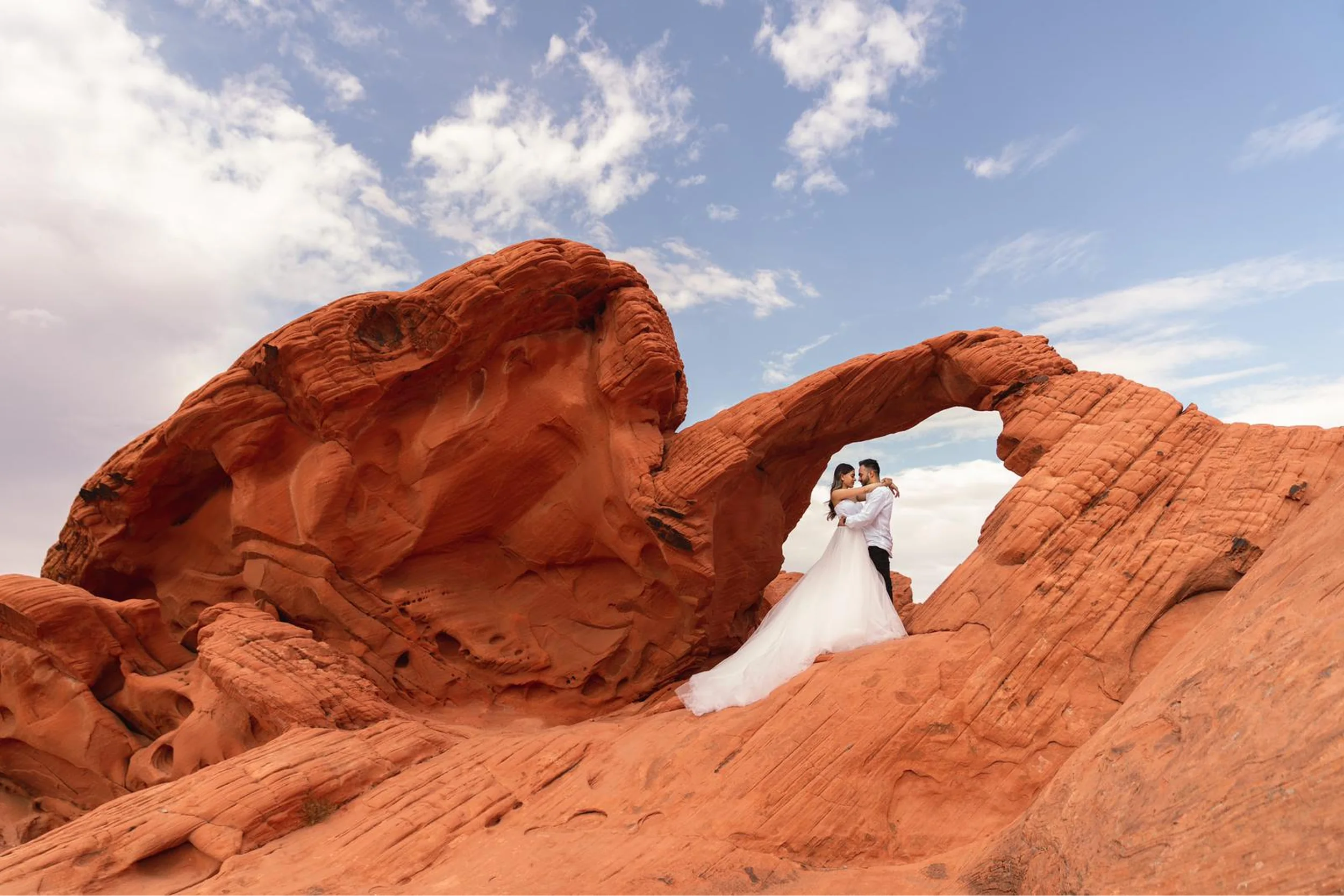 Couple in a white long-train gown embracing under a natural red rock arch during a destination photoshoot in the American Southwest