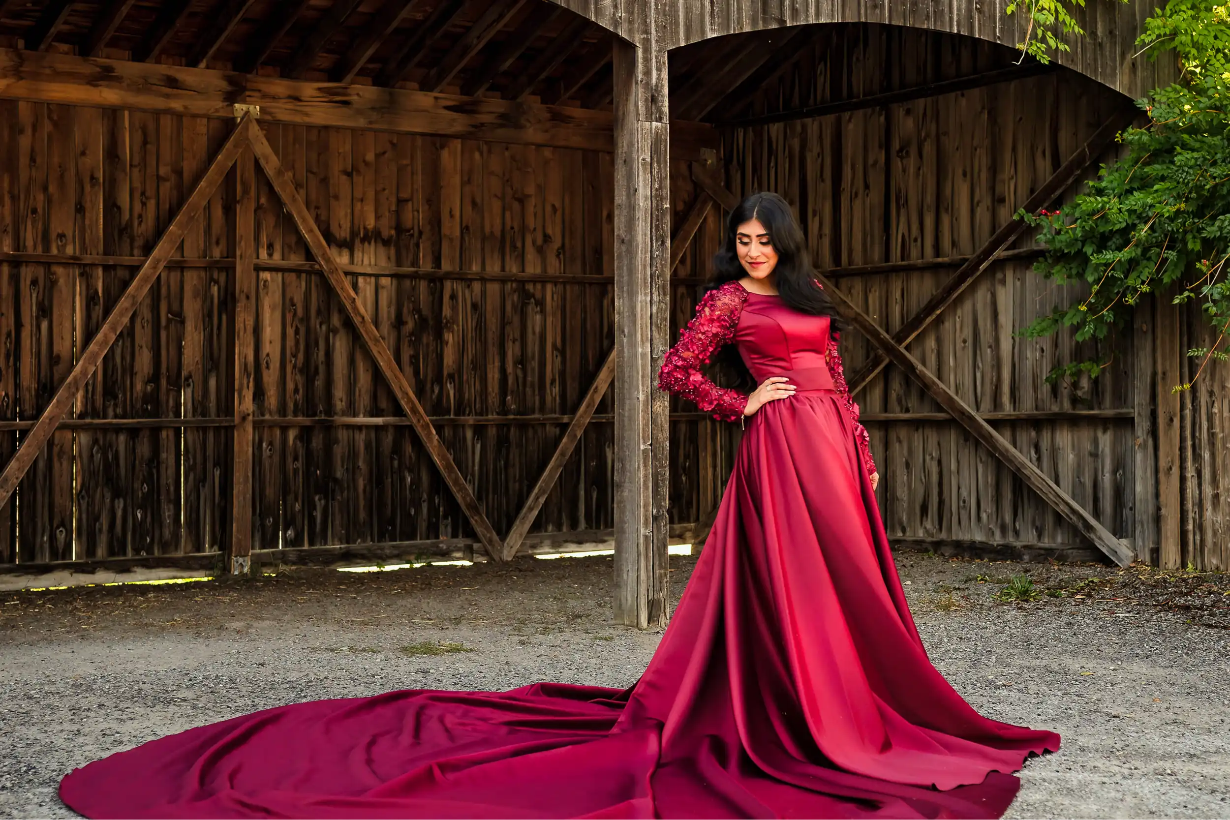 Woman in a deep burgundy long-train gown with floral lace sleeves posing in front of a rustic wooden barn during an engagement photoshoot