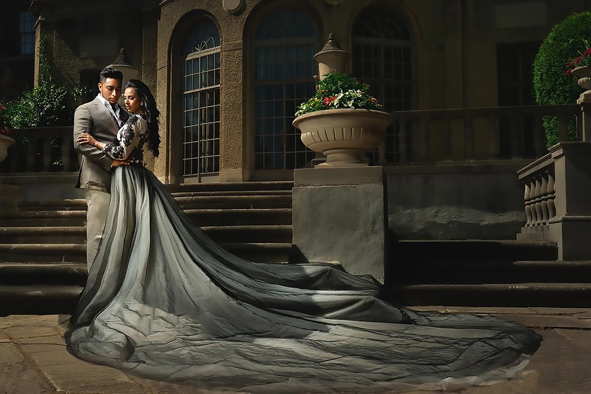 Couple embracing on mansion steps, bride's grey-black ombré long train dress flowing across the stairs
