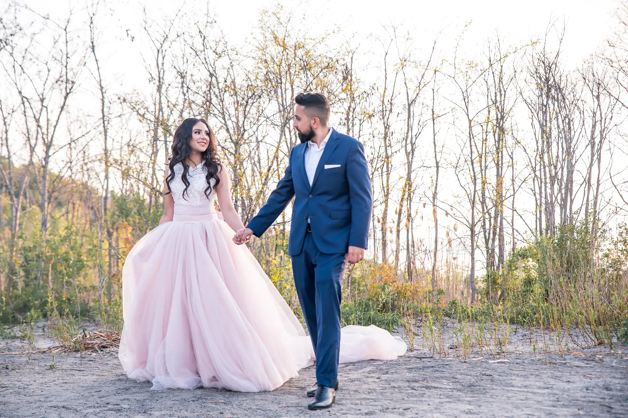 Couple walking hand-in-hand through bare trees, bride's blush pink long-train gown trailing behind on sandy path