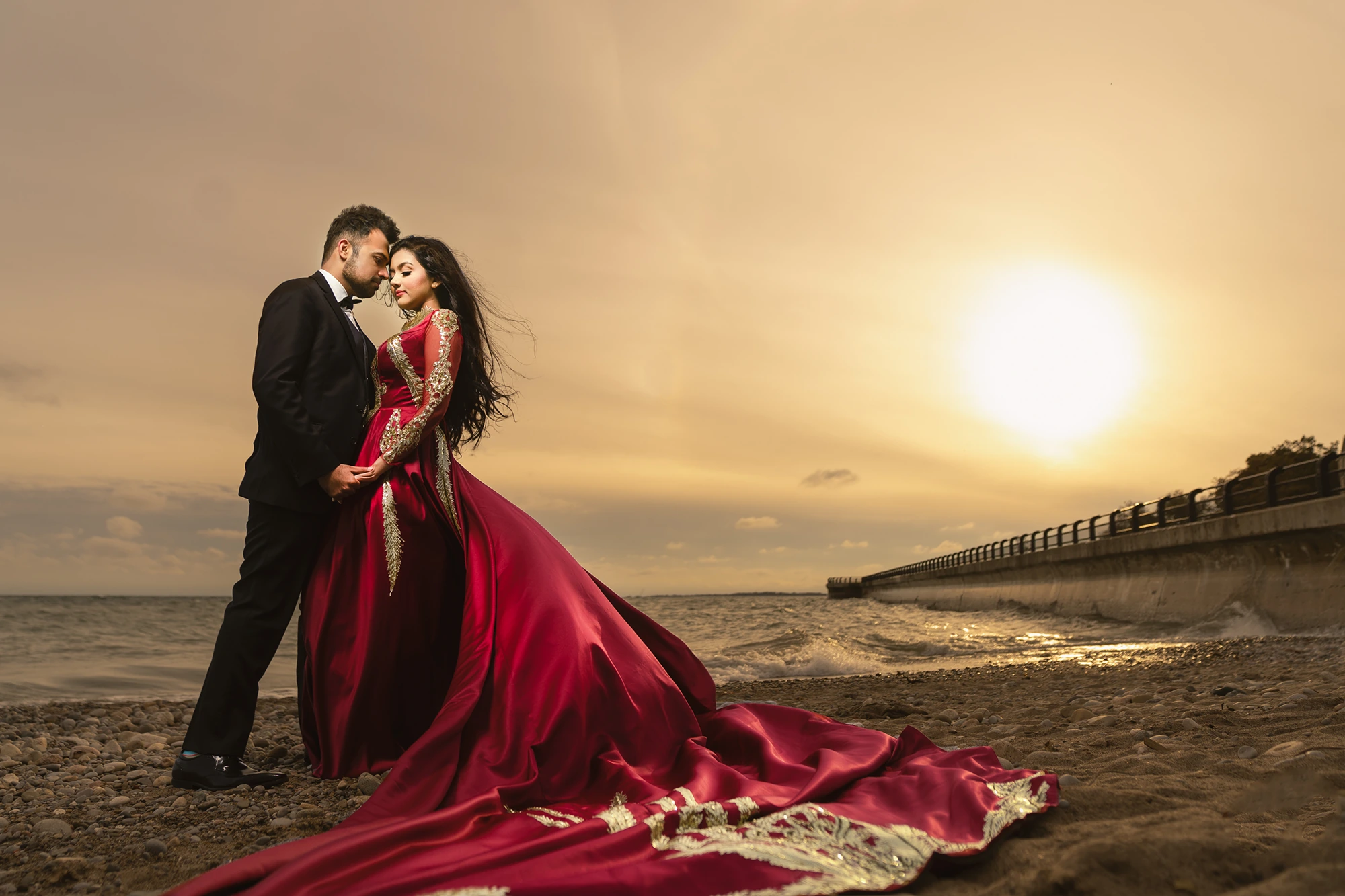 Couple at sunset on a rocky shoreline, woman in a deep red long-train gown with gold embroidery during an engagement photoshoot