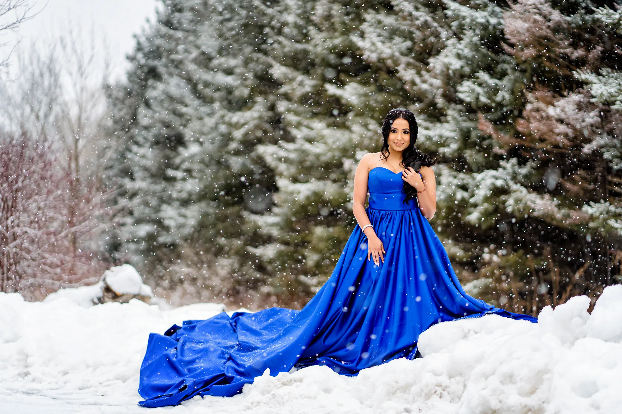 Woman in a cobalt blue long-train sweetheart gown posing in falling snow with snow-covered evergreen trees during a winter engagement photoshoot