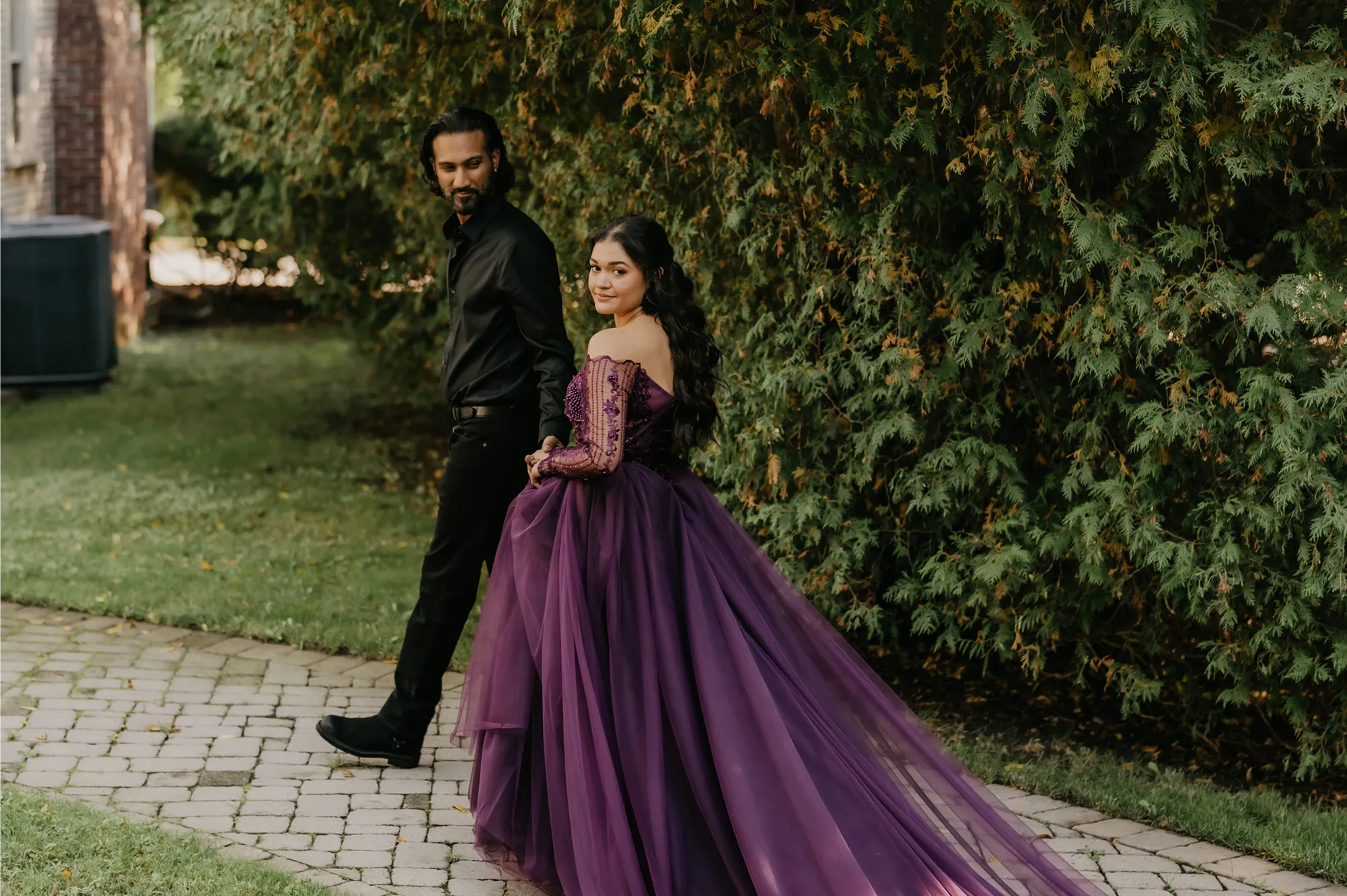 Beach or garden engagement shoot of a couple on a stone path, she wears a purple off-shoulder tulle long train gown with lace sleeves