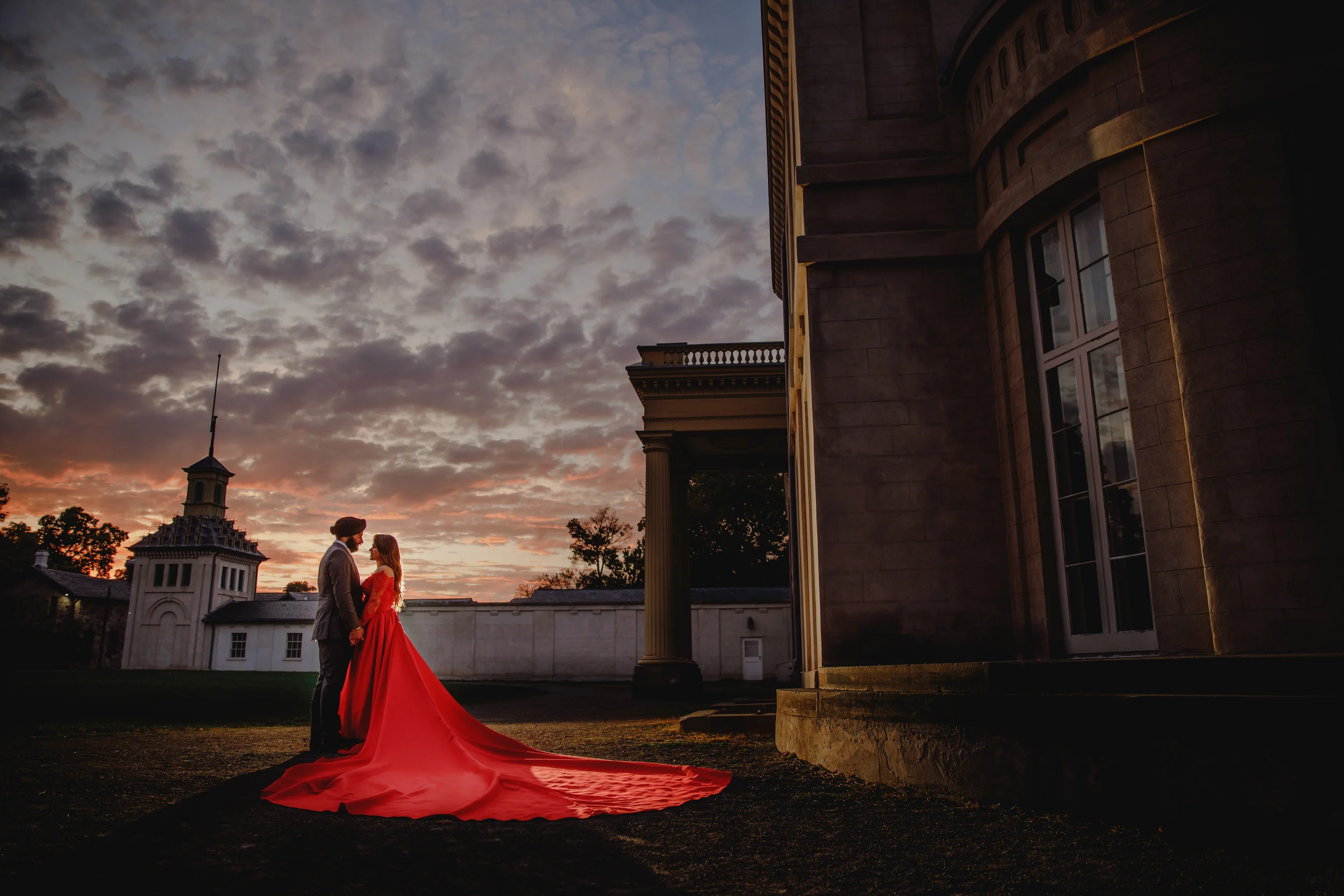 Golden hour engagement photos of a couple at sunset, she wears a red off-shoulder long train gown beside a historic stone building